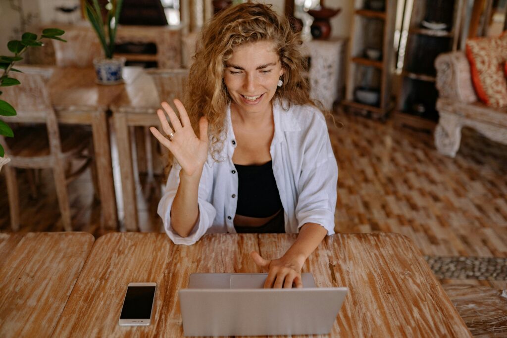Smiling woman waves during a video call on her laptop in a comfortable indoor setting.